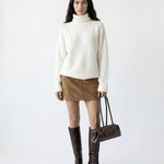 Woman wearing a white sweater, brown skirt, and brown boots holding a handbag on a white background
