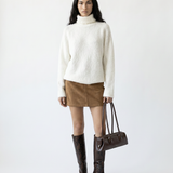 Woman wearing a white sweater, brown skirt, and brown boots holding a handbag on a white background
