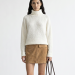 Woman wearing a white turtleneck sweater, brown suede skirt, and brown knee-high boots holding a brown handbag on a white background.