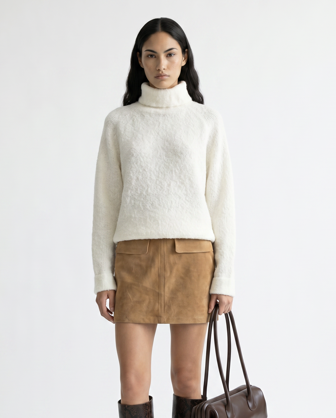 Woman wearing a white turtleneck sweater, brown suede skirt, and brown knee-high boots holding a brown handbag on a white background.