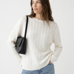 Woman wearing a white sweater and blue jeans, holding a black bag on a light gray background