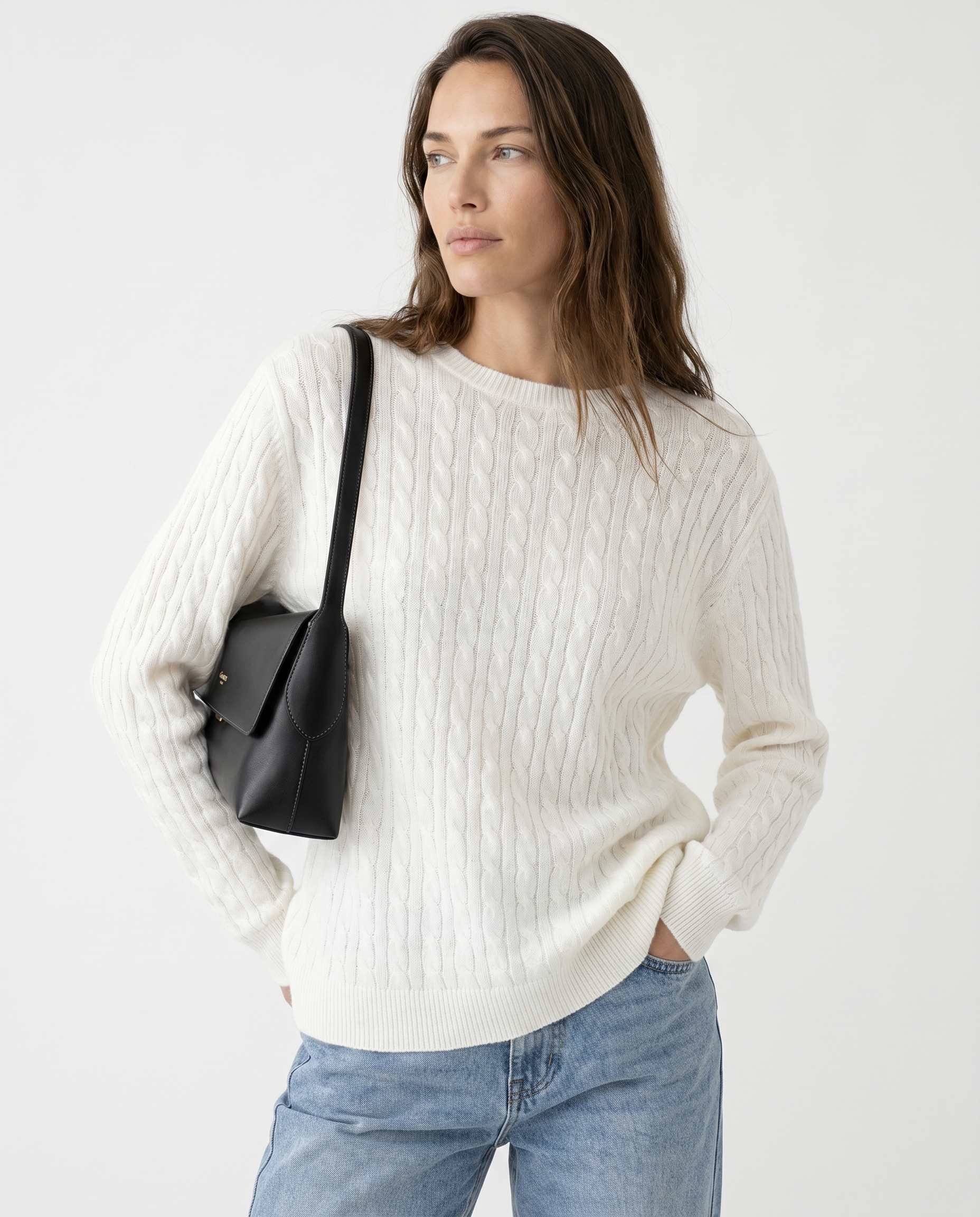 Woman wearing a white sweater and blue jeans, holding a black bag on a light gray background
