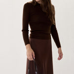 A woman wearing a dark brown mock-neck long-sleeve top and a matching dark brown midi skirt, accessorized with dark sunglasses and holding a black leather handbag against a plain off-white background.