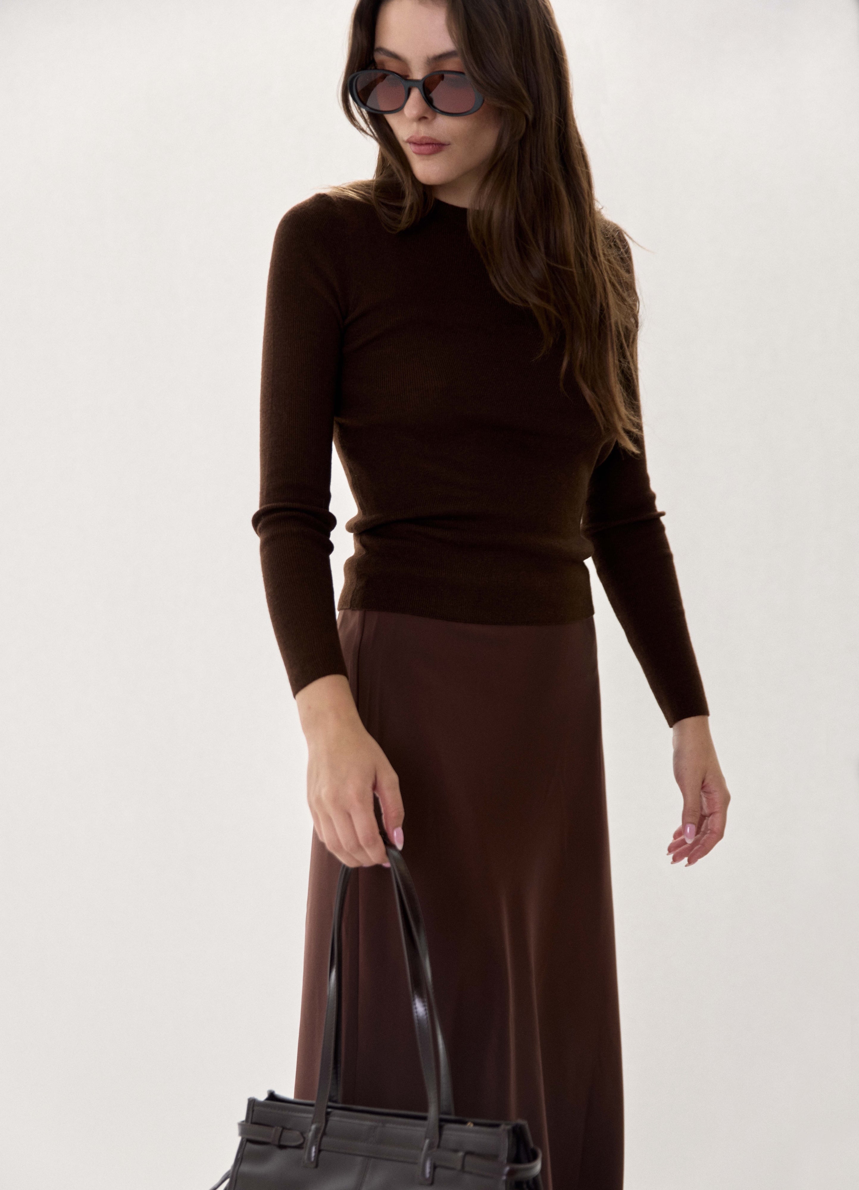 A woman wearing a dark brown mock-neck long-sleeve top and a matching dark brown midi skirt, accessorized with dark sunglasses and holding a black leather handbag against a plain off-white background.