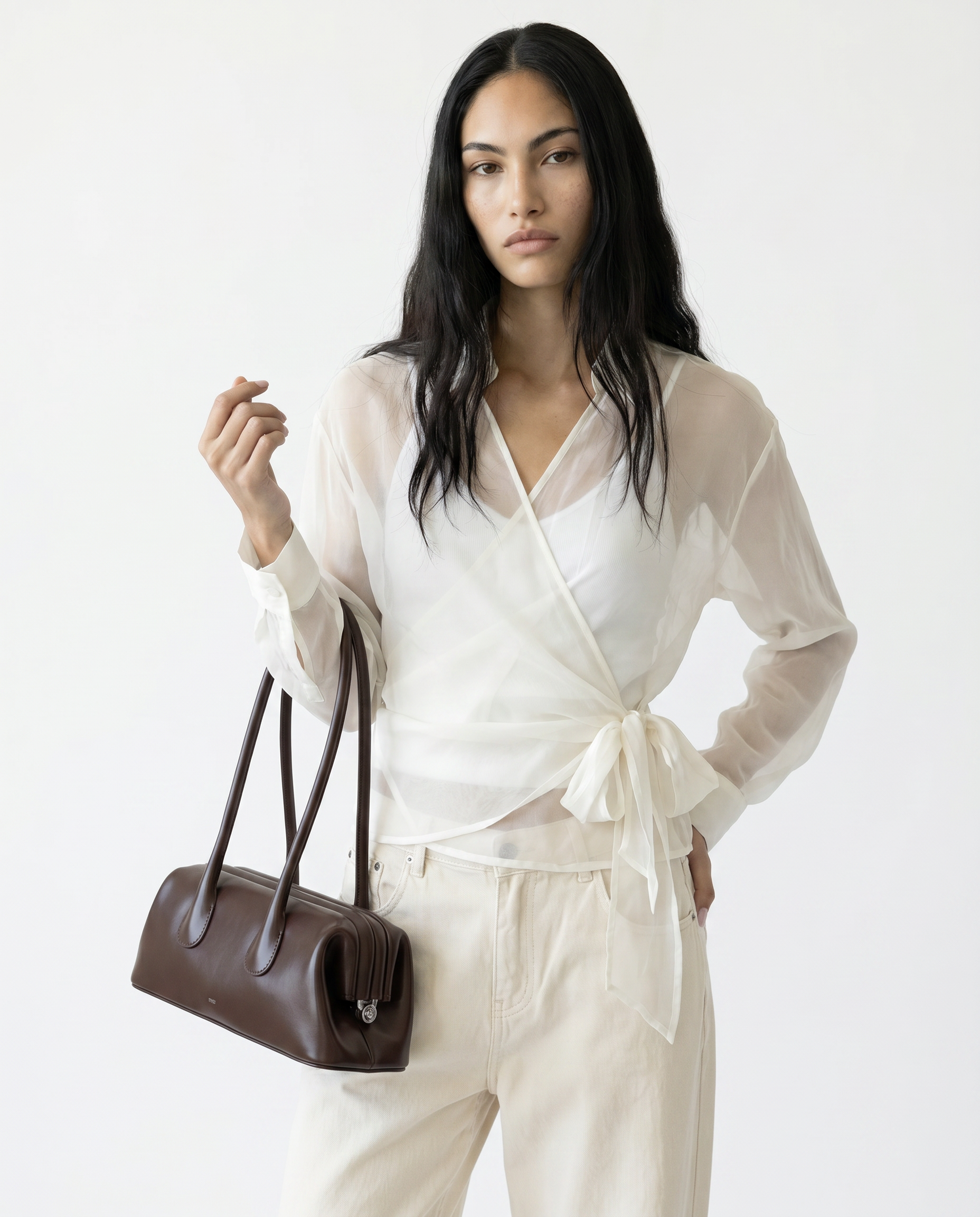 Woman in a white outfit holding a brown handbag against a white background