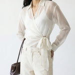 Woman wearing a white blouse with a tie waist and white pants, holding a brown handbag on a white background.