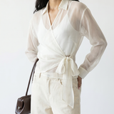 Woman wearing a white blouse with a tie waist and white pants, holding a brown handbag on a white background.