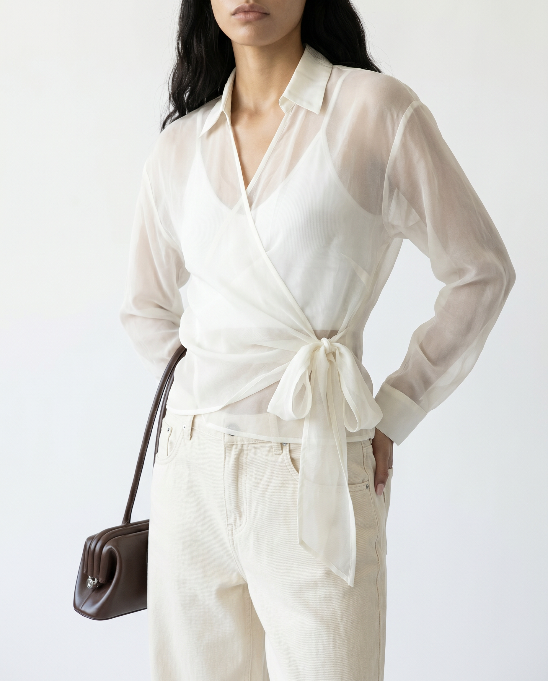 Woman wearing a white blouse with a tie waist and white pants, holding a brown handbag on a white background.