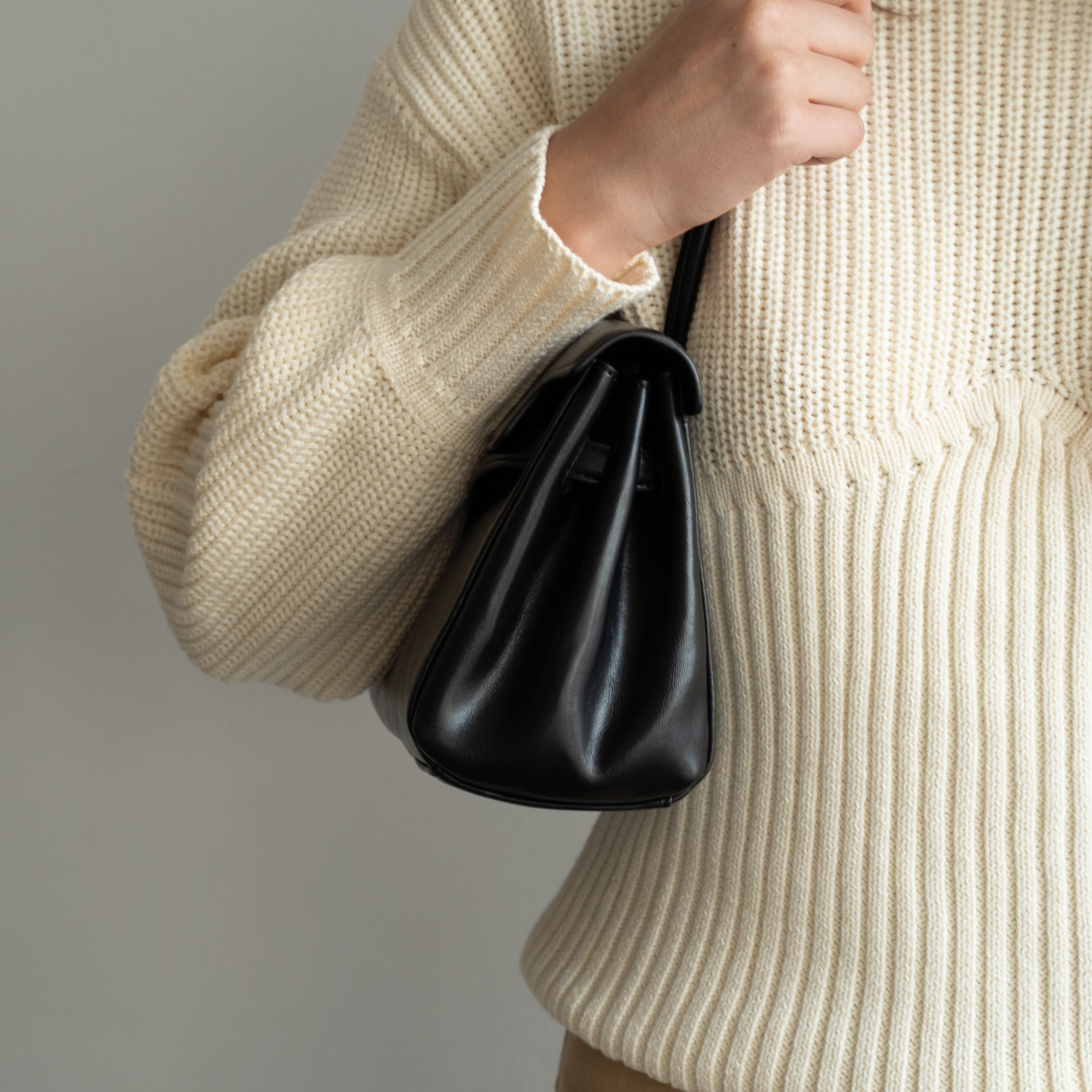 A person wearing a cream ribbed knit sweater and tan pants carries a small black leather bucket bag with a thin black shoulder strap against a plain light gray background.
