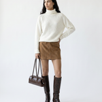 Woman wearing a white sweater, brown skirt, and knee-high boots holding a brown handbag on a white background