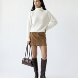 Woman wearing a white sweater, brown skirt, and knee-high boots holding a brown handbag on a white background