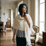 A woman wearing a cream-colored fringed shawl or poncho over a black dress, standing in a classic, elegant room with wooden floors and large windows.