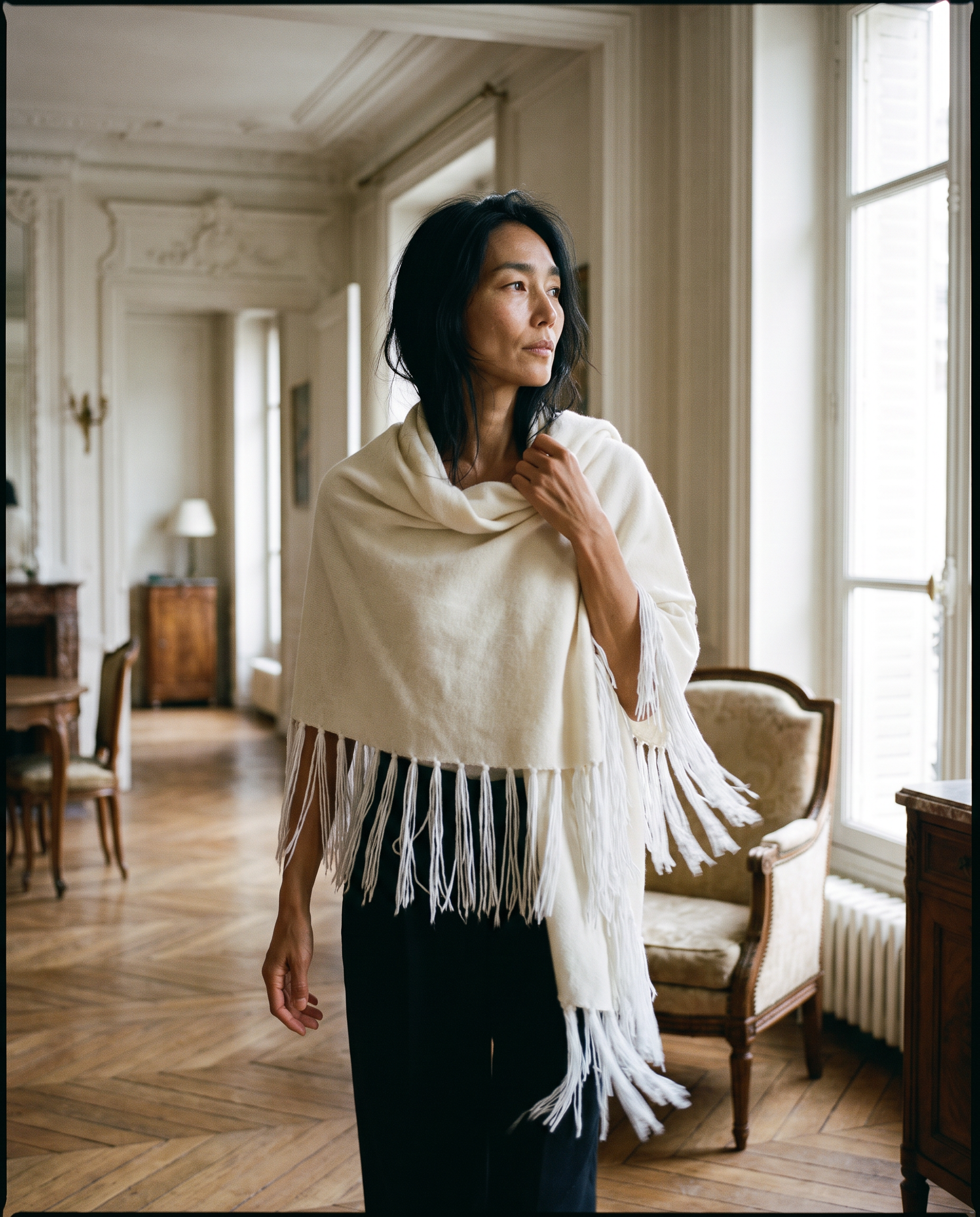A woman wearing a cream-colored fringed shawl or poncho over a black dress, standing in a classic, elegant room with wooden floors and large windows.