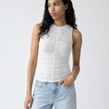 A woman modeling a white sleeveless ruched top with a high mock neck, paired with light-wash blue jeans, against a plain light gray studio background.