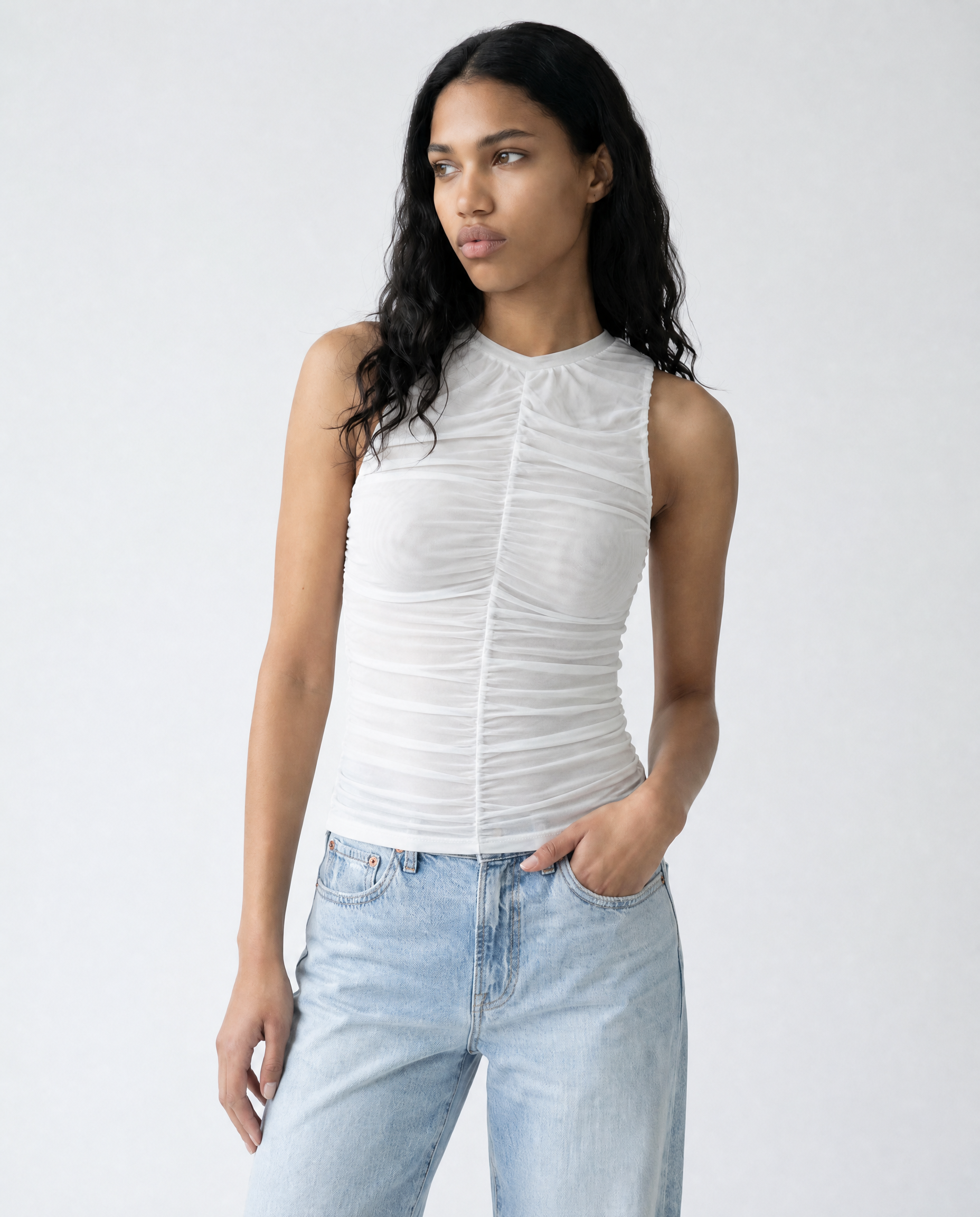 A woman modeling a white sleeveless ruched top with a high mock neck, paired with light-wash blue jeans, against a plain light gray studio background.