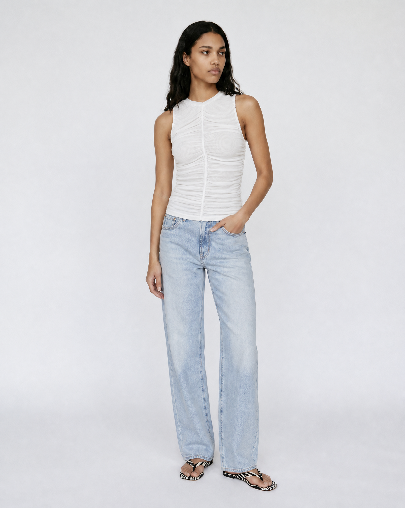 A model stands against a light gray studio background wearing a white sleeveless ruched top, light-wash wide-leg jeans, and leopard-print sandals.