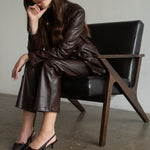 A woman wearing a dark brown faux-leather jacket and matching wide-leg pants sits on a black leather chair with a wooden frame in a minimalist room.