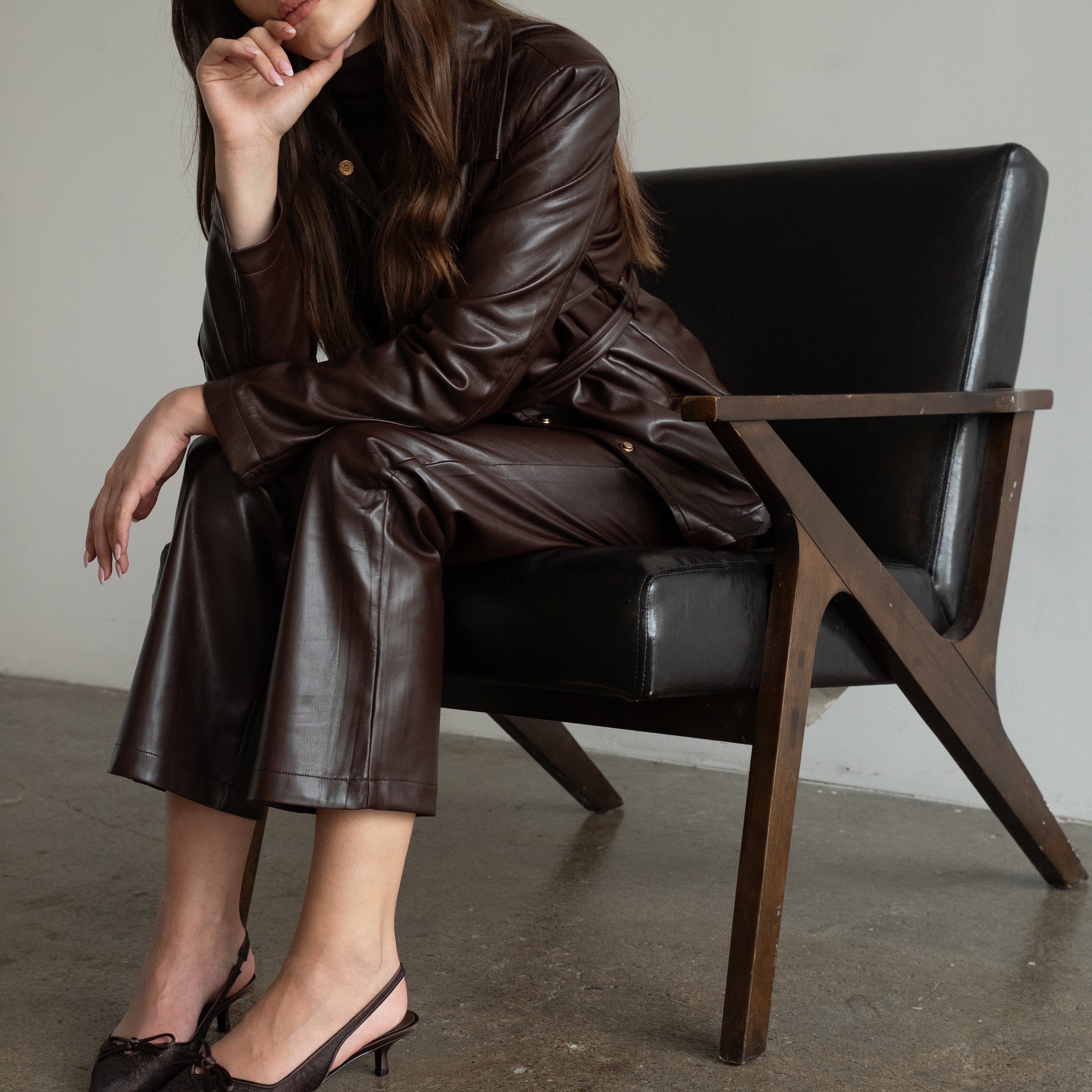 A woman wearing a dark brown faux-leather jacket and matching wide-leg pants sits on a black leather chair with a wooden frame in a minimalist room.