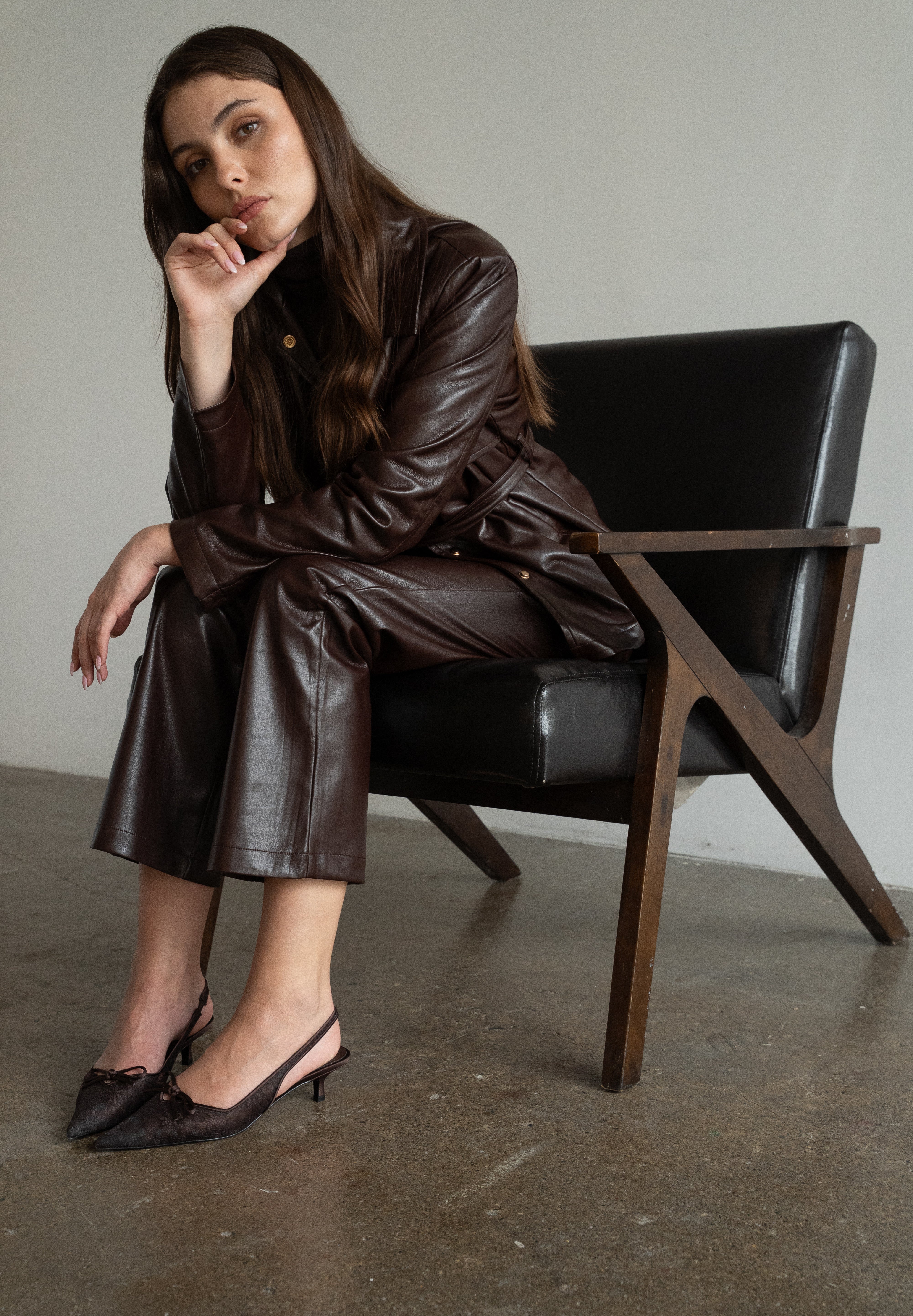 A woman wearing a dark brown faux-leather jacket and matching wide-leg pants sits on a black leather chair with a wooden frame in a minimalist room.