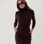 A woman modeling a dark brown ribbed turtleneck sweater and matching dark brown midi skirt against a plain light gray studio background.