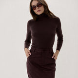 A woman modeling a dark brown ribbed turtleneck sweater and matching dark brown midi skirt against a plain light gray studio background.