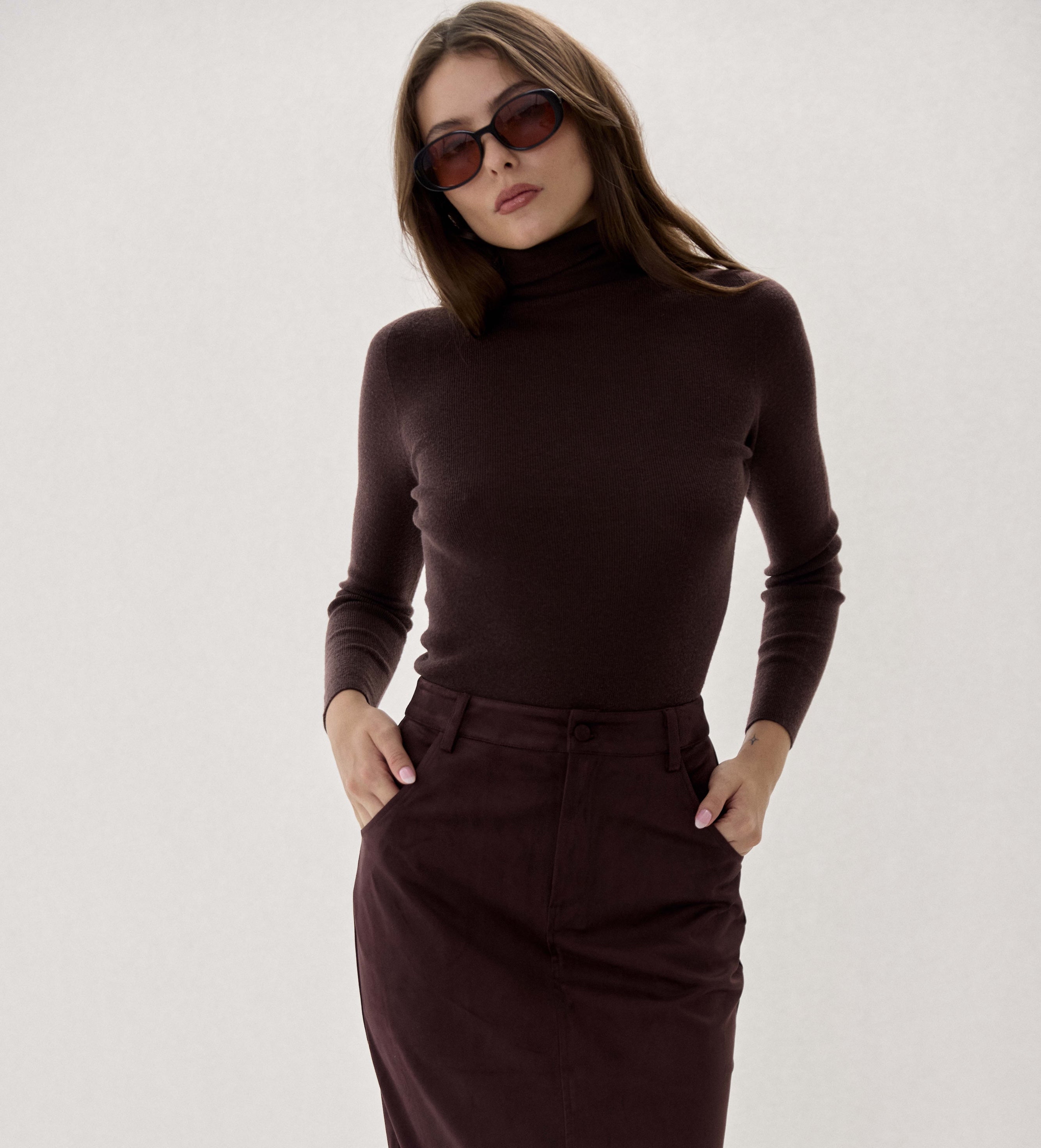 A woman modeling a dark brown ribbed turtleneck sweater and matching dark brown midi skirt against a plain light gray studio background.