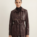 A model wearing a dark brown leather belted jacket over a black turtleneck, paired with matching dark brown leather trousers, against a light beige studio background.
