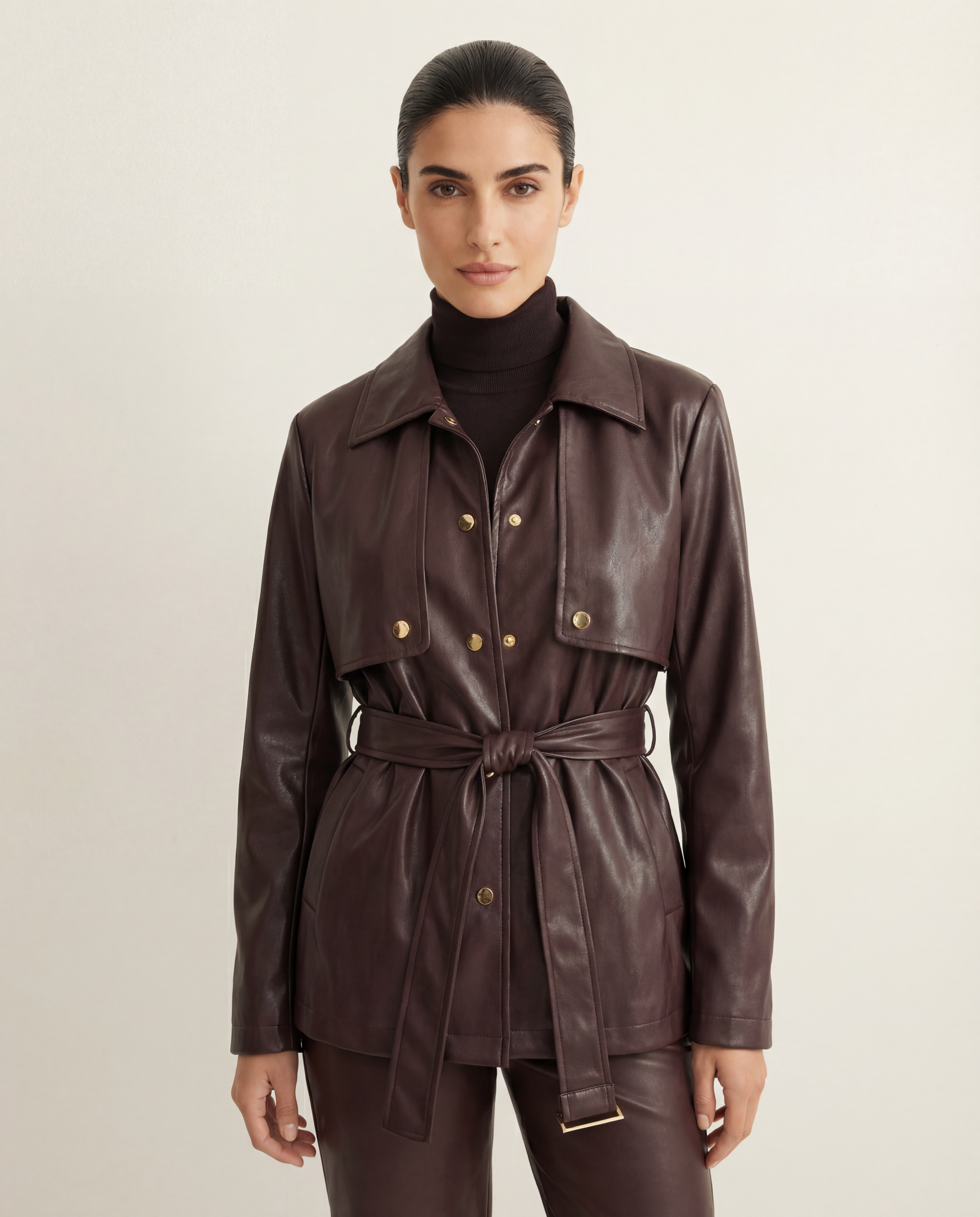A model wearing a dark brown leather belted jacket over a black turtleneck, paired with matching dark brown leather trousers, against a light beige studio background.