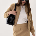 A woman models a tan two-piece jacket and skirt set, styled with a white top, black sunglasses, and a black croc-embossed shoulder bag against a plain light background.