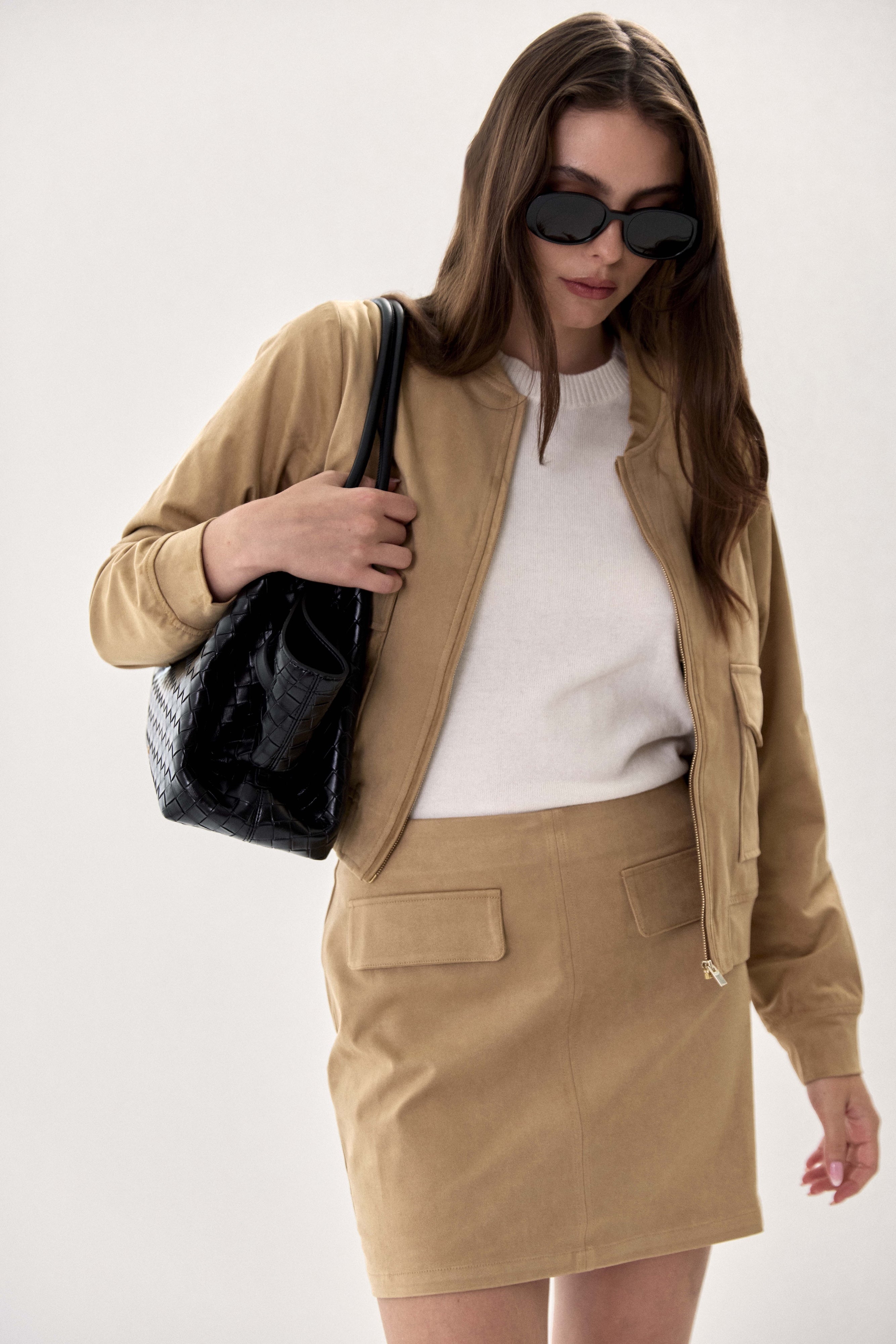 A woman models a tan two-piece jacket and skirt set, styled with a white top, black sunglasses, and a black croc-embossed shoulder bag against a plain light background.
