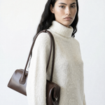 Woman wearing a white sweater holding a brown handbag on a light gray background