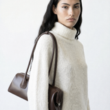 Woman wearing a white sweater holding a brown handbag on a light gray background