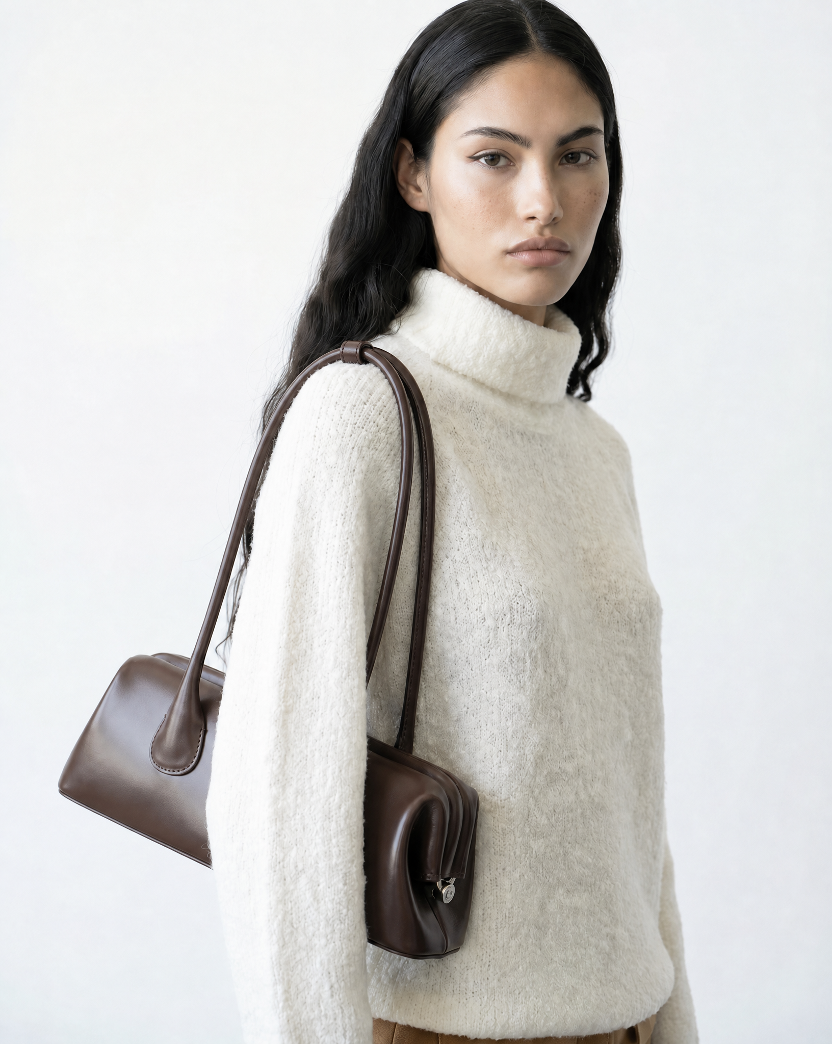 Woman wearing a white sweater holding a brown handbag on a light gray background