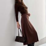 A woman stands against a white wall wearing a dark brown long-sleeve top and matching brown midi skirt, paired with black pointed-toe ankle boots and holding a structured dark brown handbag.