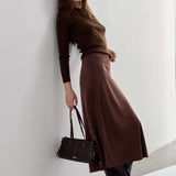 A woman stands against a white wall wearing a dark brown long-sleeve top and matching brown midi skirt, paired with black pointed-toe ankle boots and holding a structured dark brown handbag.