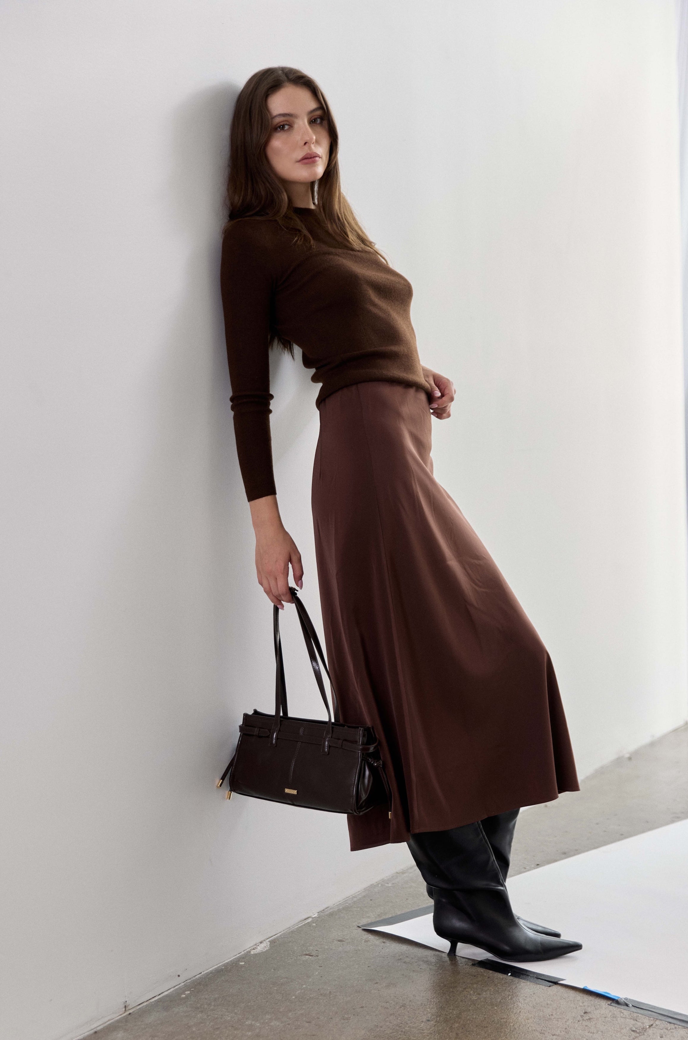 A woman stands against a white wall wearing a dark brown long-sleeve top and matching brown midi skirt, paired with black pointed-toe ankle boots and holding a structured dark brown handbag.