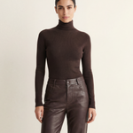 A model stands against a plain light beige studio background wearing a dark brown ribbed turtleneck sweater and matching dark brown leather trousers.
