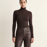 A model stands against a plain light beige studio background wearing a dark brown ribbed turtleneck sweater and matching dark brown leather trousers.