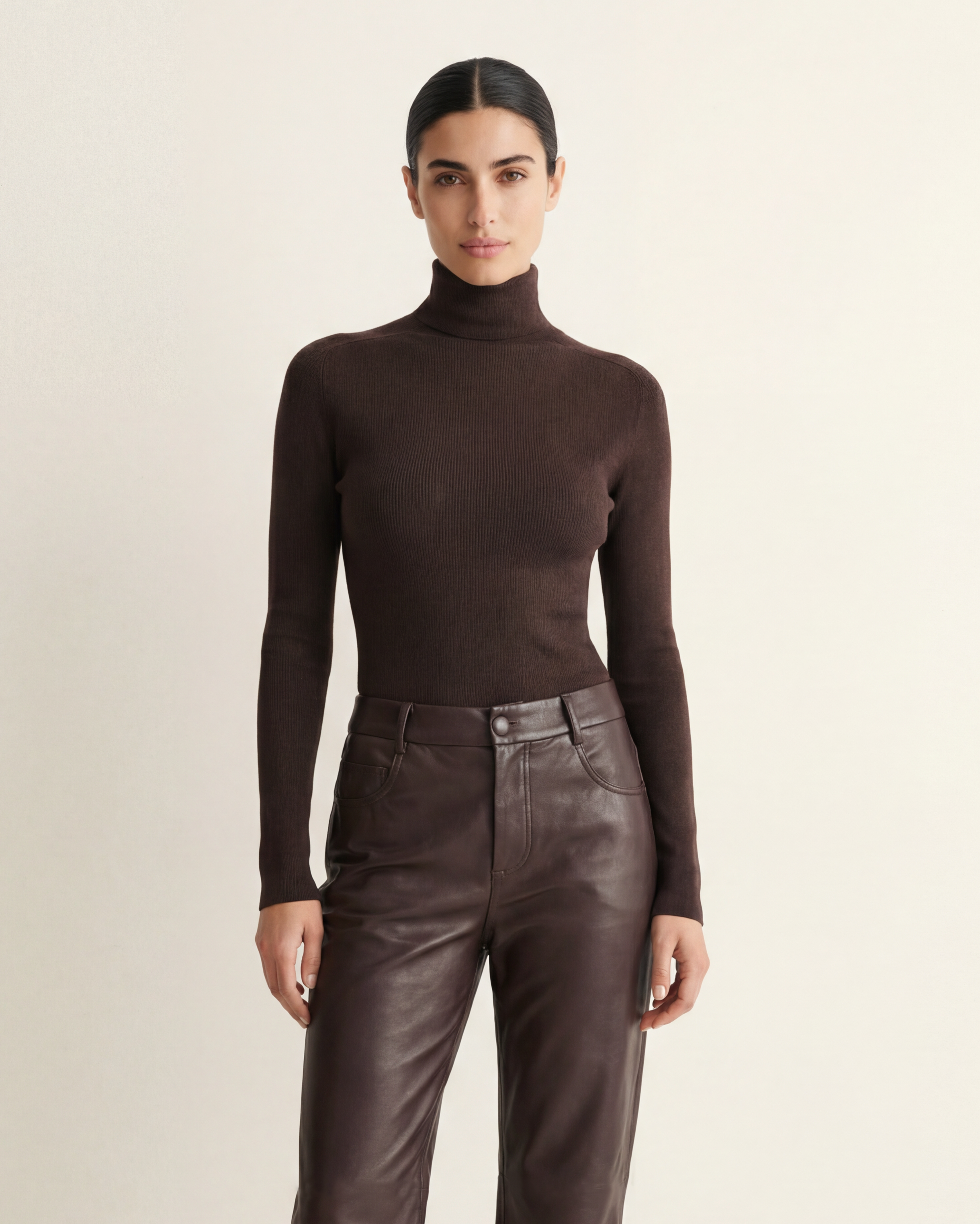 A model stands against a plain light beige studio background wearing a dark brown ribbed turtleneck sweater and matching dark brown leather trousers.